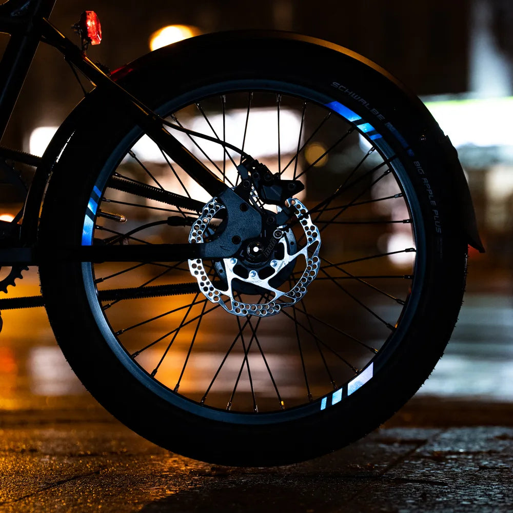Reflektierender Aufkleber „Racer“ in Farbvariante „Weltraum“ in Größe 20mm auf schwarzer Fahrrad Felge im Dunkeln mit verschwommenen Lichtern im Hintergrund. von REFLECTIVE Berlin.