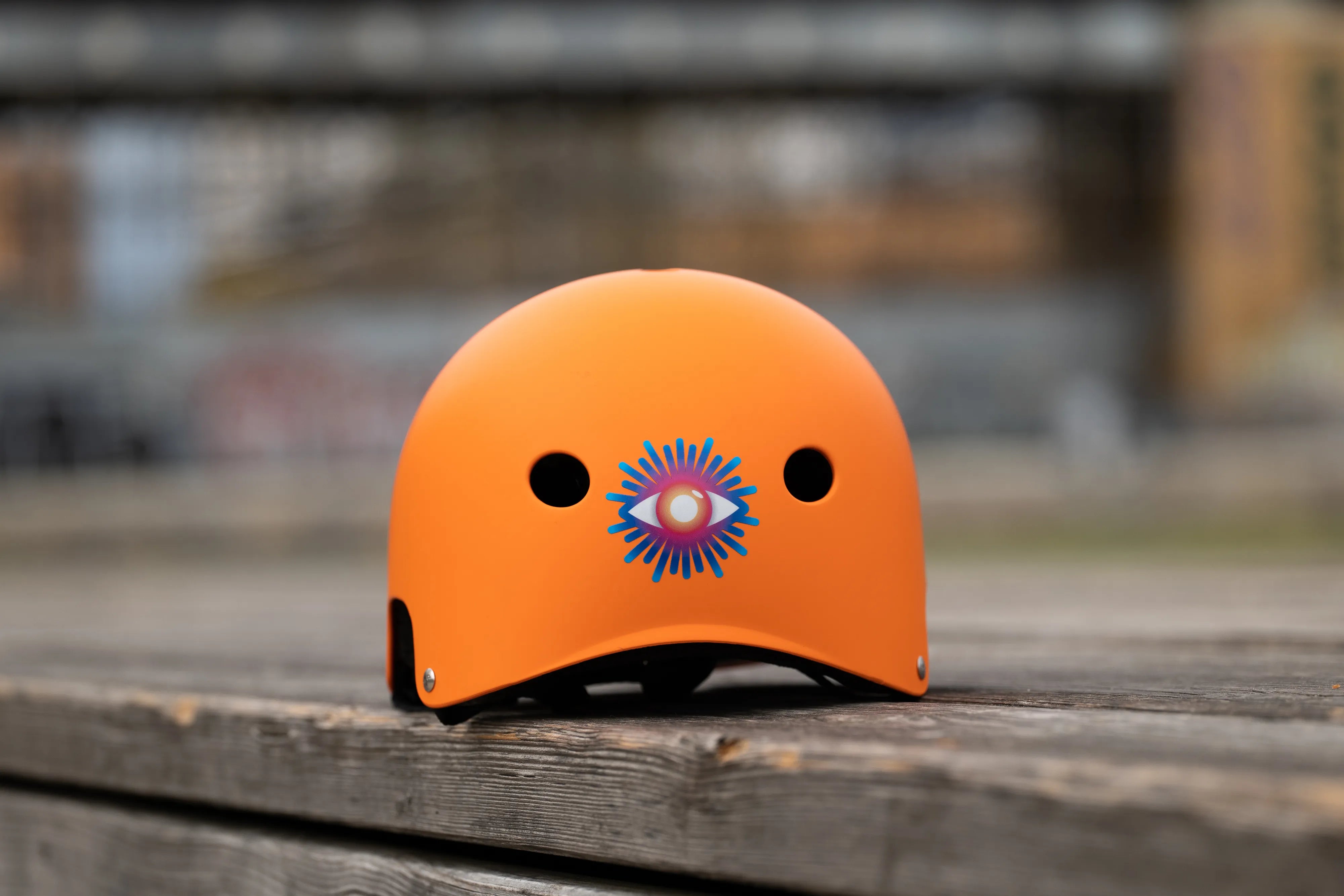 Reflektierender Aufkleber „Auge“ in Farbvariante „Regenbogen“ auf orangem Fahrradhelm Rückansicht vor unscharfem Hintergrund. Von REFLECTIVE Berlin.