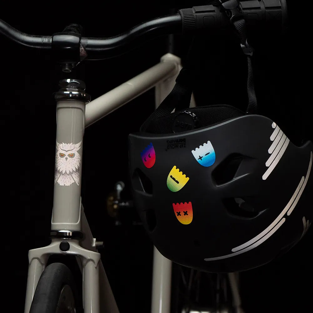 Reflektierender Aufkleber „Geister“ in Farbvariante „Regenbogen“ auf einem schwarzen Fahrradhelm der am Lenker eines Fahrrads hängt. Von REFLECTIVE Berlin.