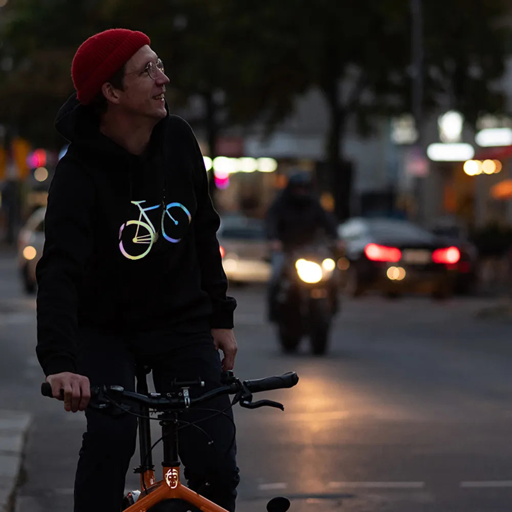Person mit roter Mütze sitzt auf einem orangen BULLIT Lastenrad und trägt den reflektierenden Hoodie Fahrrad in Schwarz von REFLECTIVE Berlin bei Dämmerung mit sichtbarer Reflexion.