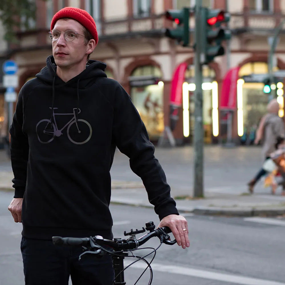 Person mit roter Mütze trägt den reflektierenden Hoodie Fahrrad in Schwarz von REFLECTIVE Berlin in einer urbanen Berliner Straßenszene bei Tageslicht.