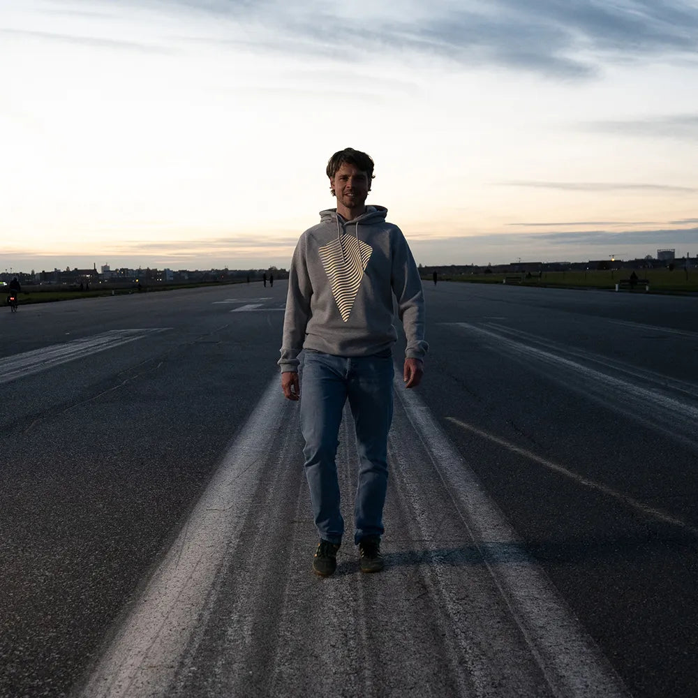 Person trägt den reflektierenden Hoodie Dune in Grau von REFLECTIVE Berlin auf dem Tempelhofer Feld während der blauen Stunde. Das Design reflektiert.