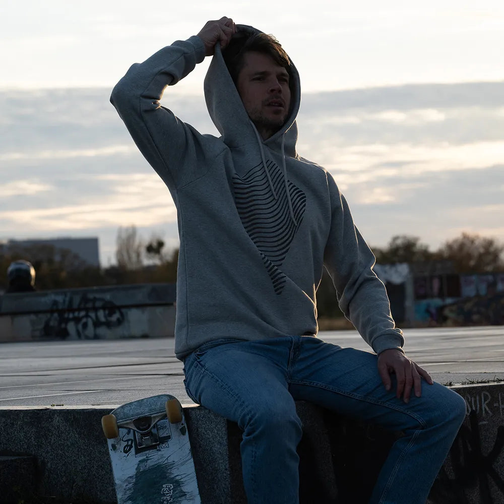 Person trägt den reflektierenden Hoodie Dune in Grau von REFLECTIVE Berlin und sitzt neben einem Skateboard auf dem Tempelhofer Feld bei Tageslicht.