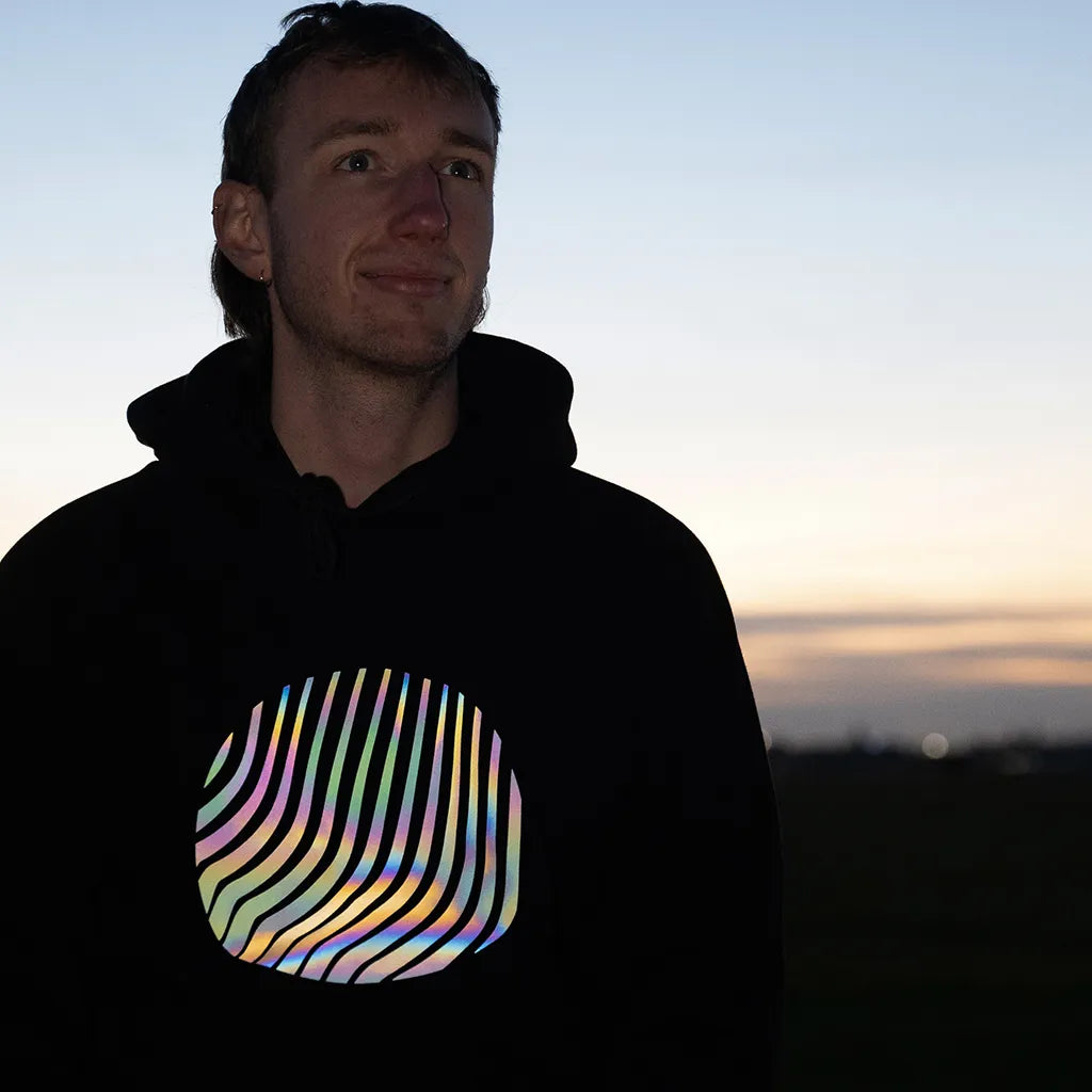 Person trägt den reflektierenden Hoodie Sunset in Schwarz mit Regenbogen-Reflexion von REFLECTIVE Berlin auf dem Tempelhofer Feld während der blauen Stunde. Die Reflexion ist stark sichtbar.