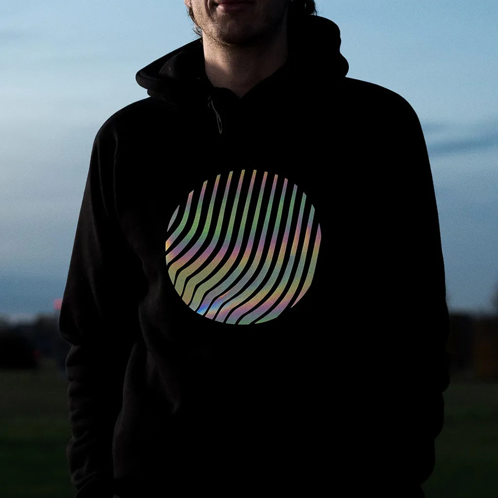 Person trägt den reflektierenden Hoodie Sunset in Schwarz mit Regenbogen-Reflexion von REFLECTIVE Berlin auf dem Tempelhofer Feld in der Dämmerung.