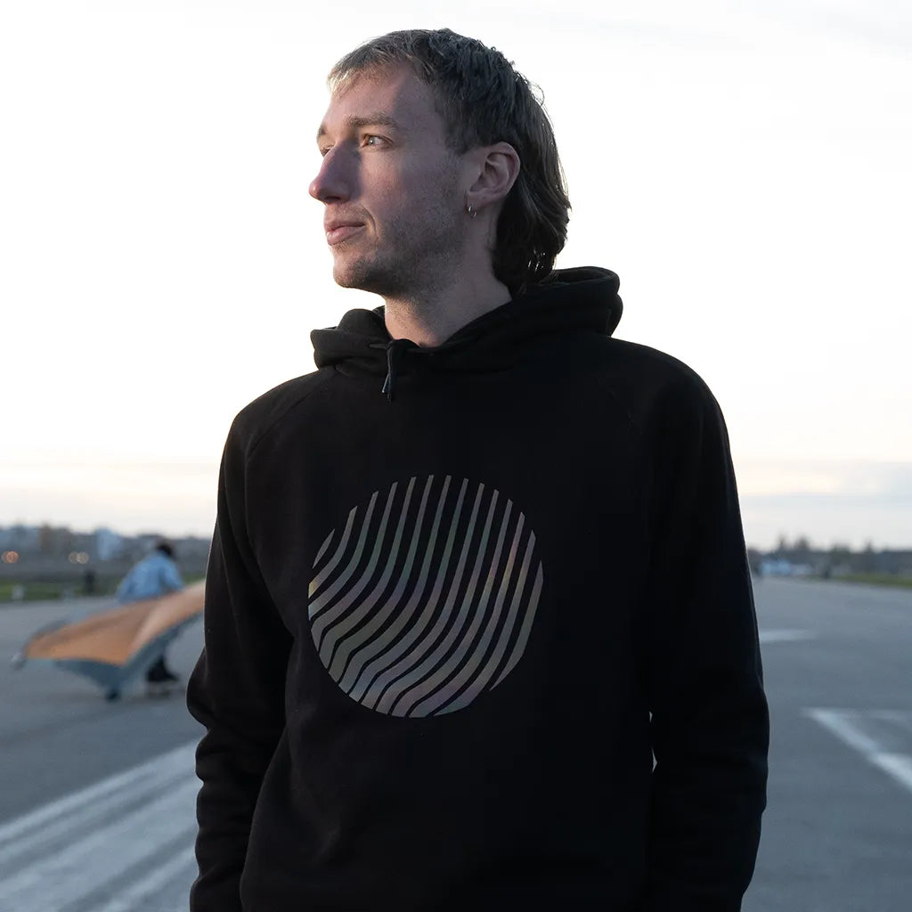 Person trägt den reflektierenden Hoodie Sunset in Schwarz mit Regenbogen-Reflexion von REFLECTIVE Berlin bei Tageslicht.