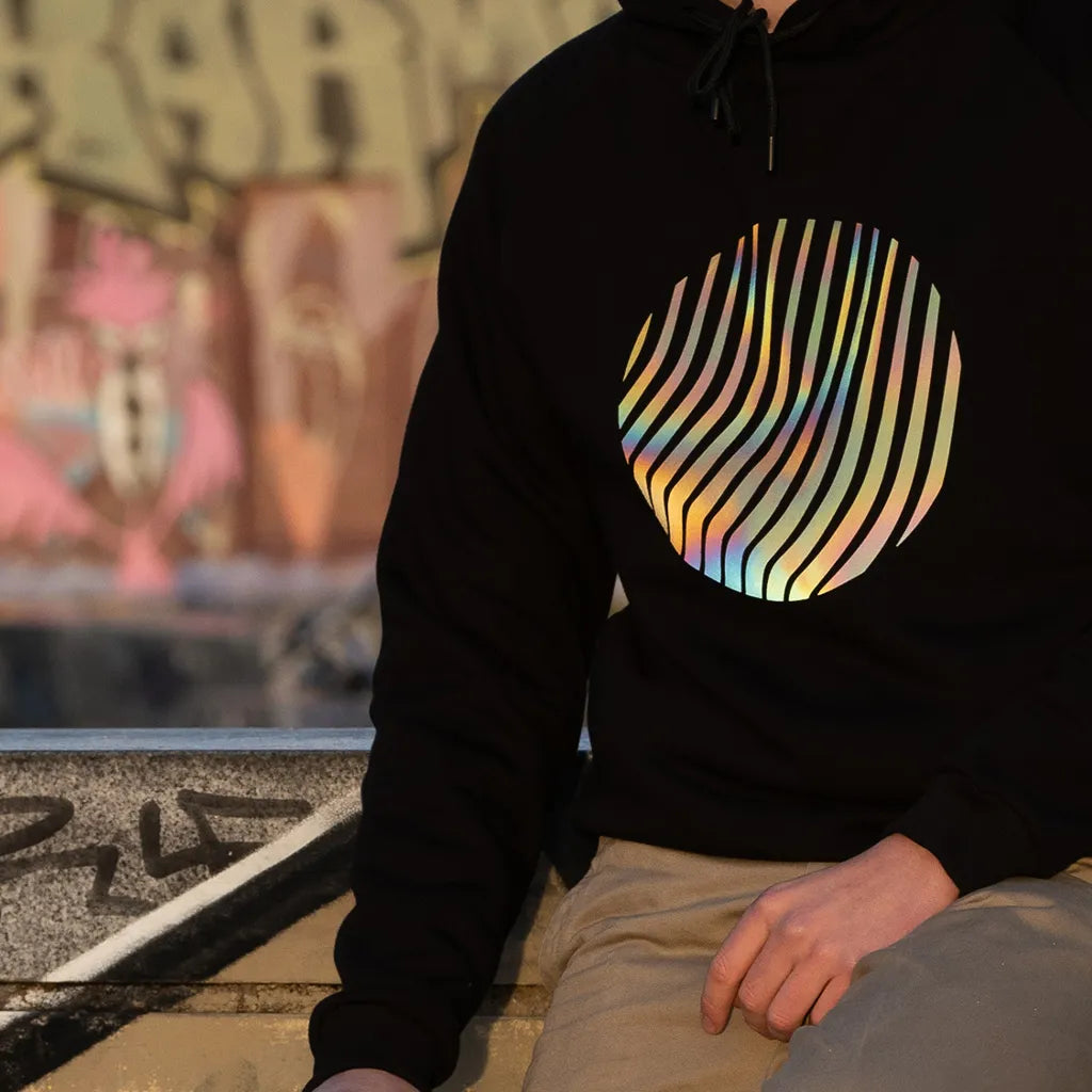 Person trägt den reflektierenden Hoodie Sunset in Schwarz mit Regenbogen-Reflexion von REFLECTIVE Berlin bei Tageslicht. Die Sonne reflektiert auf dem Print.