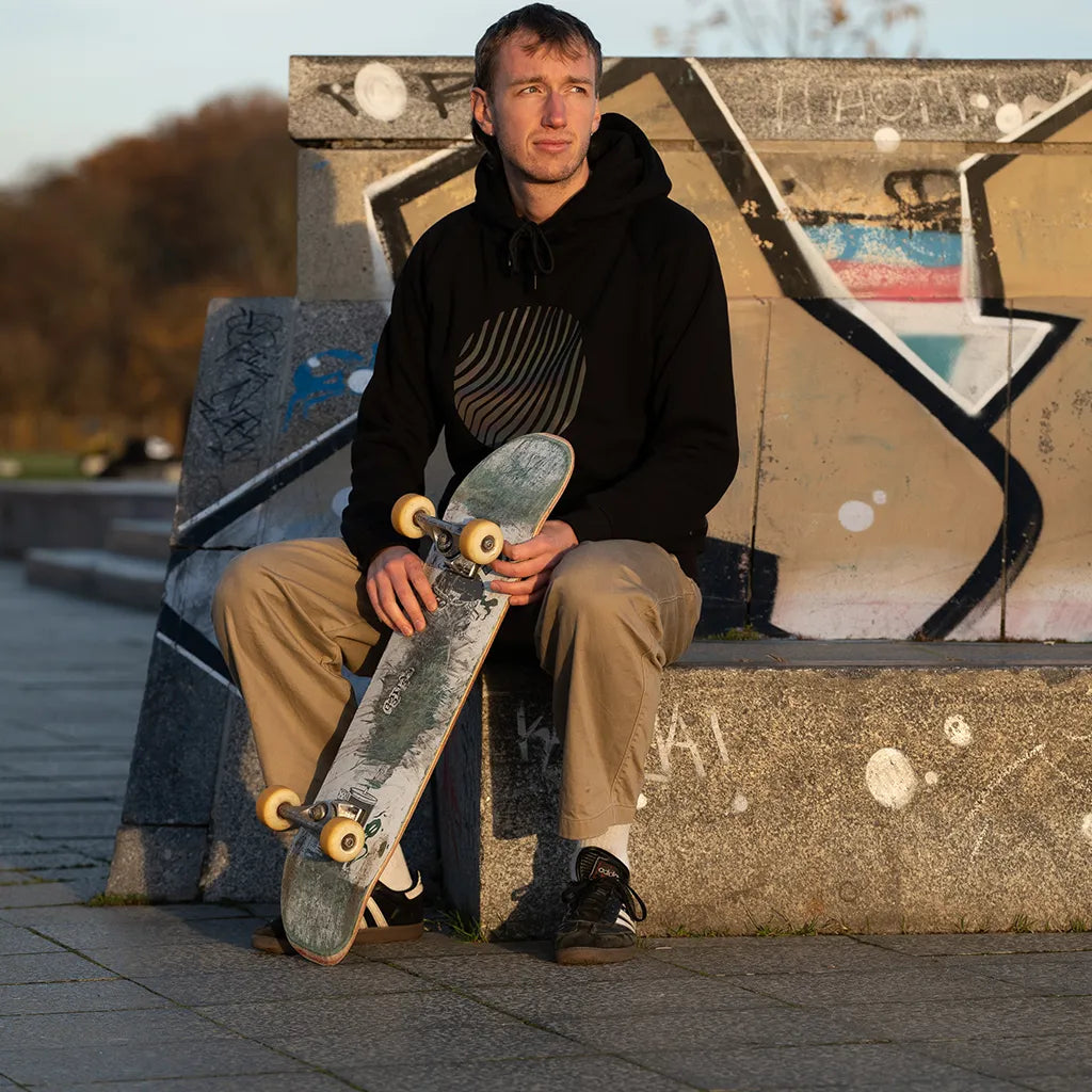 Person mit Skateboard trägt den reflektierenden Hoodie Sunset in Schwarz mit Regenbogen-Reflexion von REFLECTIVE Berlin bei Tageslicht.