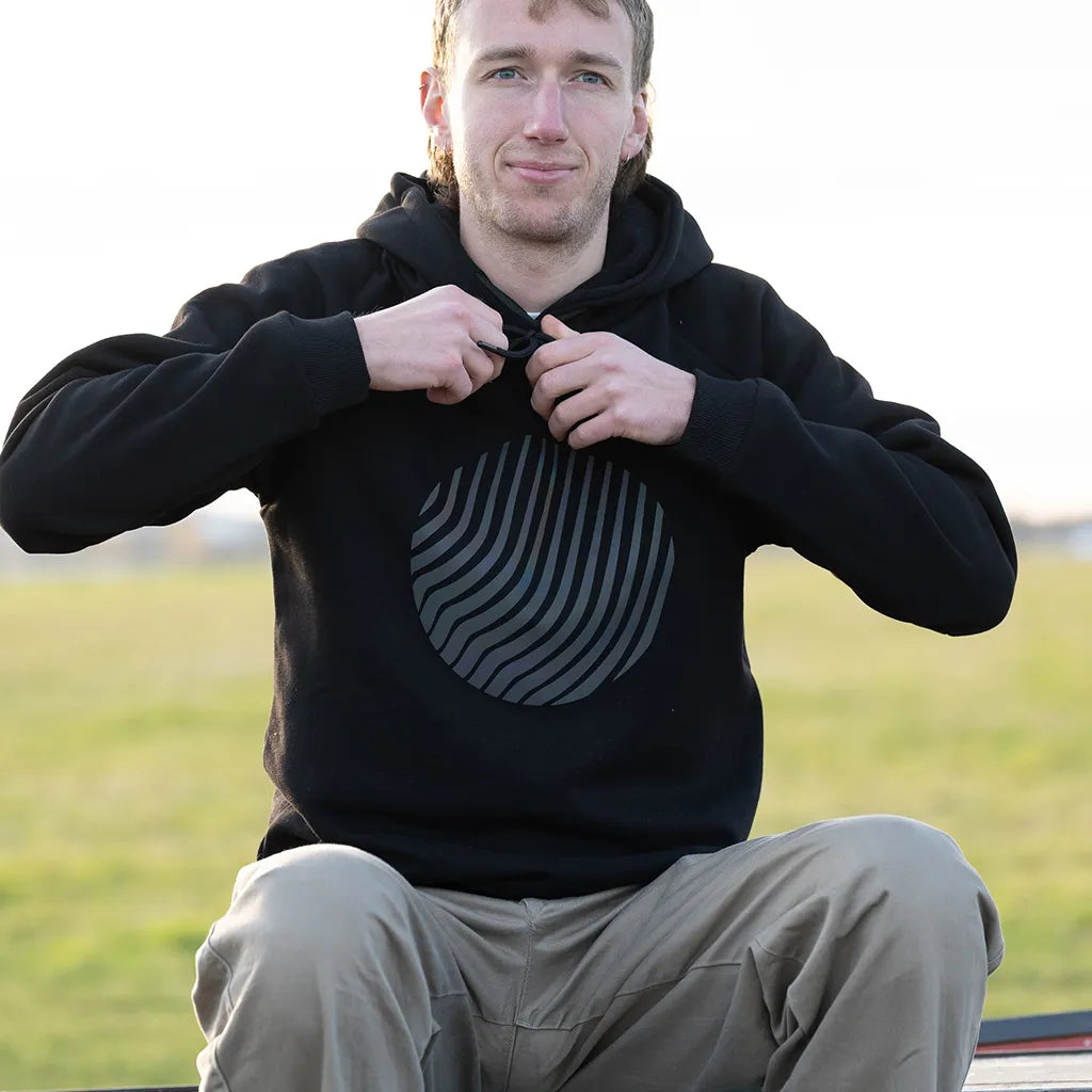 Sitzende Person trägt den reflektierenden Hoodie Sunset in Schwarz mit Regenbogen-Reflexion von REFLECTIVE Berlin auf dem Tempelhofer Feld bei Tageslicht.