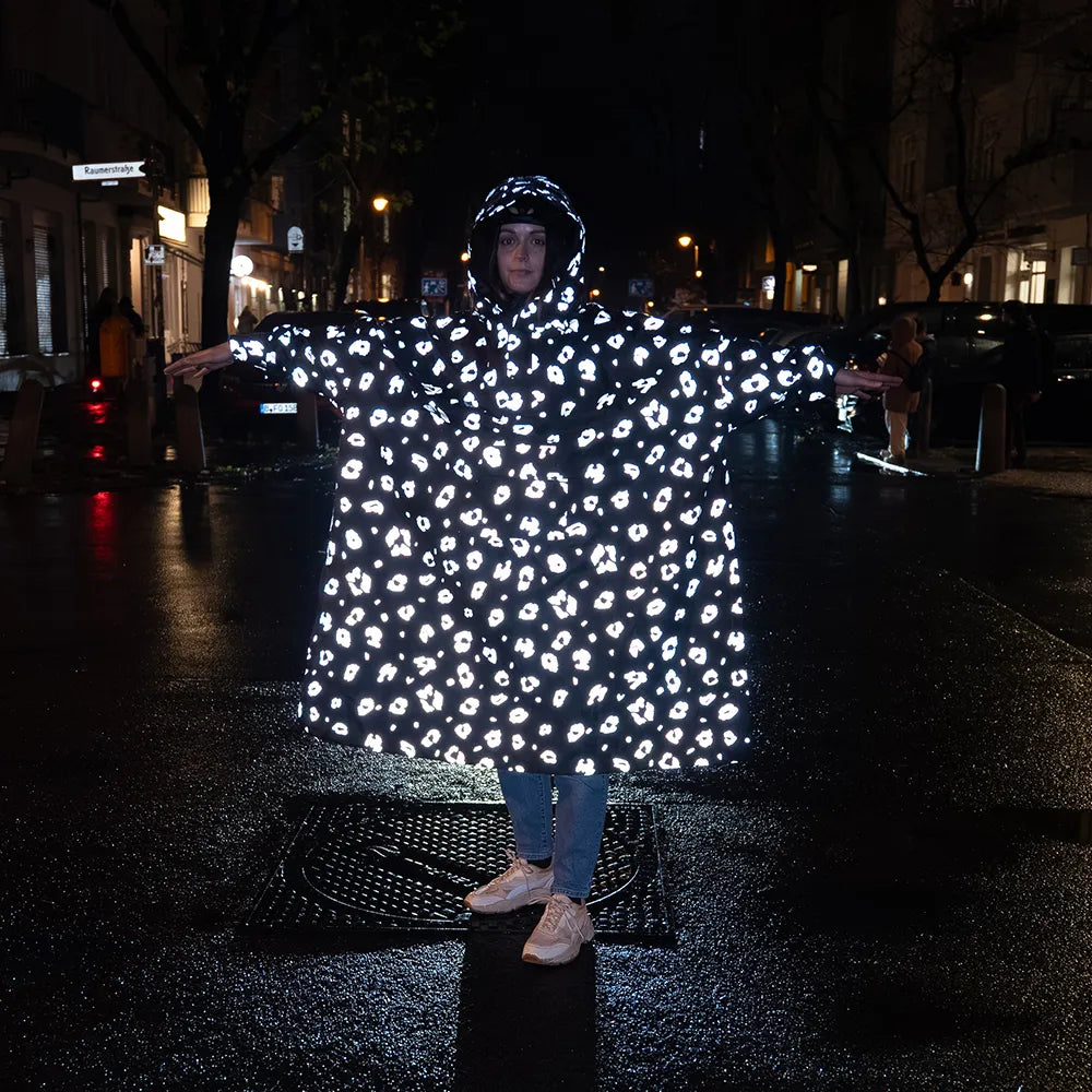reflektierender Fahrrad Regenponcho in Farbvariante „Leo“ getragen von einer Person, die im Regen auf einer Straße steht. Das Bild wurde bei Dunkelheit mit Blitzlicht aufgenommen, um die Reflexion des Ponchos hervorzuheben. Kollaboration von Bomance und REFLECTIVE Berlin.