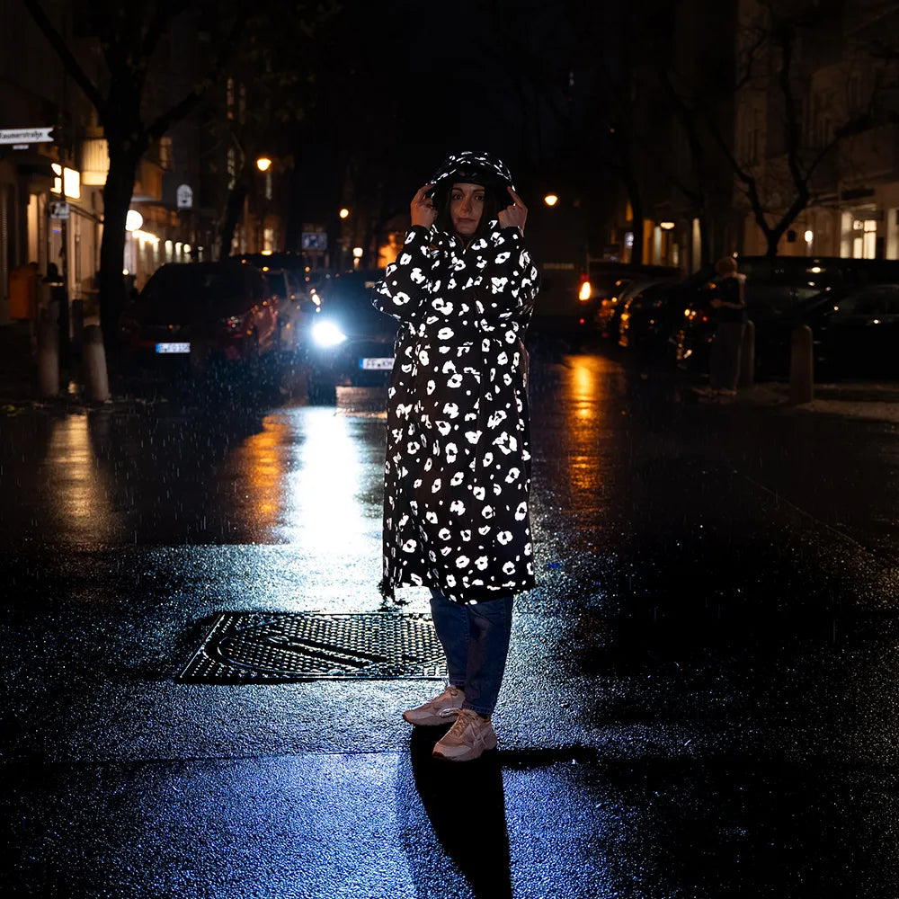 reflektierender Fahrrad Regenponcho in Farbvariante „Leo“, getragen von einer Person, die die Kapuze festhält und von einem Auto angestrahlt wird. Das Bild wurde bei Dunkelheit mit Blitzlicht aufgenommen, um die Reflexion des Ponchos hervorzuheben. Kollaboration von Bomance und REFLECTIVE Berlin.