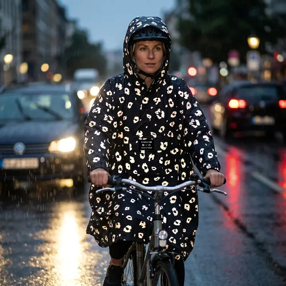 reflektierender Fahrrad Regenponcho in Farbvariante „Leo“, getragen von einer Person, die bei Regenwetter auf ihrem Fahrrad sitzt und von Autos hinter ihr angestrahlt wird. Das Bild wurde bei Dunkelheit mit Blitzlicht aufgenommen, um die Reflexion des Ponchos hervorzuheben. Kollaboration von Bomance und REFLECTIVE Berlin.