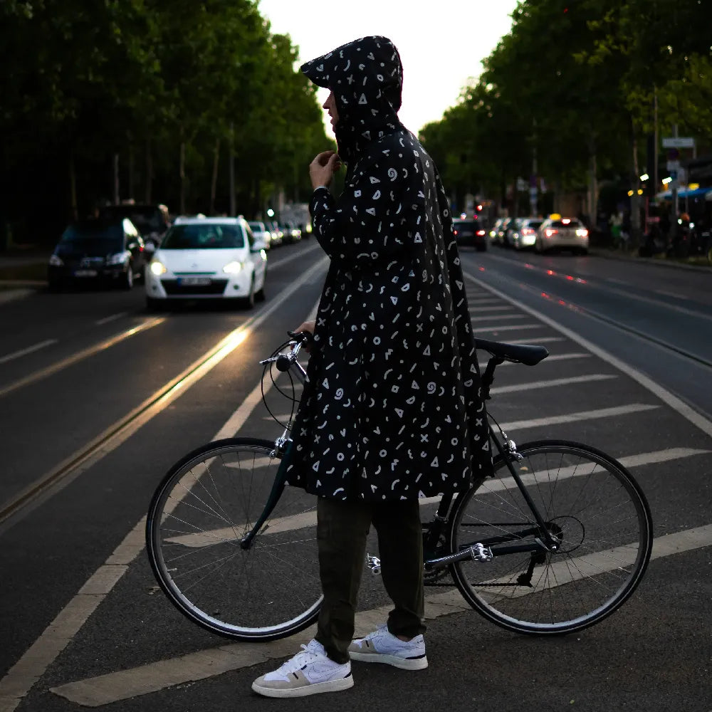 reflektierender Fahrrad Regenponcho in Farbvariante „Doodle“ getragen von einer Person, die einen Fahrradhelm trägt und auf dem Mittelstreifen einer Straße steht. Kollaboration von Bomance und REFLECTIVE Berlin.
