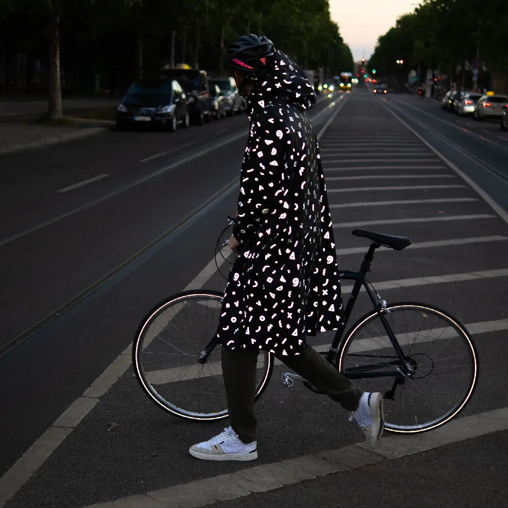 stark reflektierender Fahrrad Regenponcho in Farbvariante „Doodle“ getragen von einer Person im Profil, die einen Fahrradhelm trägt und mit einem Fahrrad über eine Straße geht. Kollaboration von Bomance und REFLECTIVE Berlin.