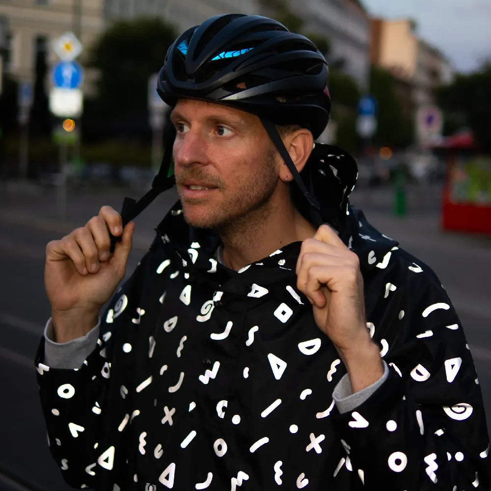 stark reflektierender Fahrrad Regenponcho in Farbvariante „Doodle“ getragen von einer Person, die einen Fahrradhelm trägt und über eineStraße geht. Die Doodle Elemente reflektieren stark. Kollaboration von Bomance und REFLECTIVE Berlin.