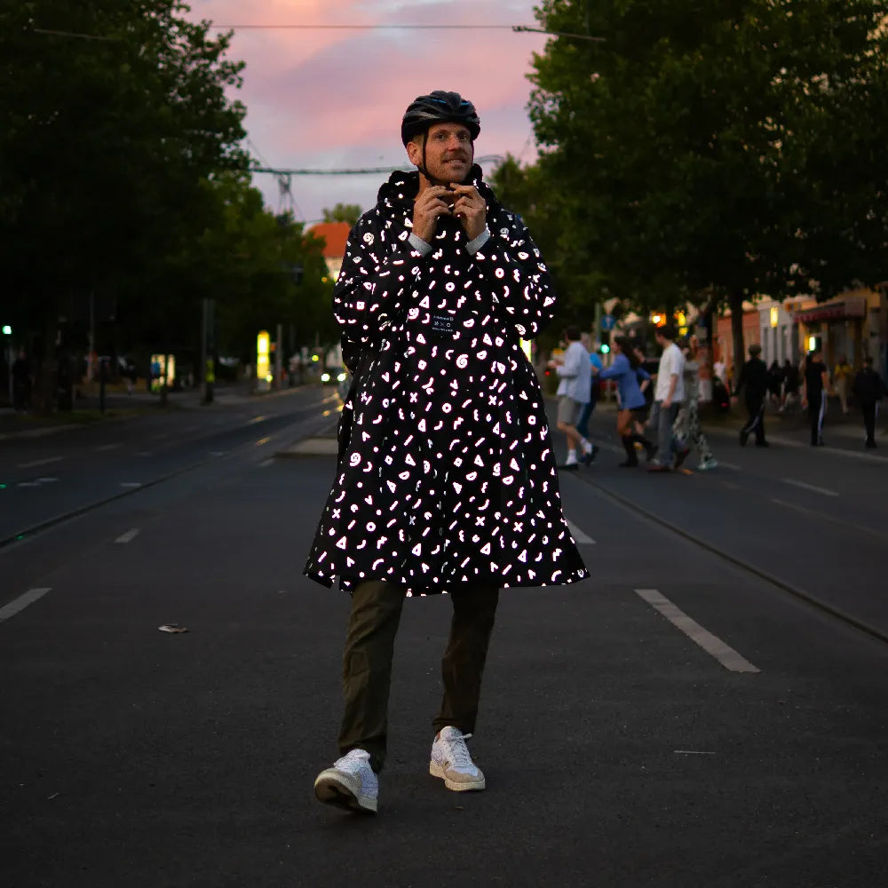 stark reflektierender Fahrrad Regenponcho in Farbvariante „Doodle“ getragen von einer Person, die einen Fahrradhelm trägt und auf einer Straße steht. Kollaboration von Bomance und REFLECTIVE Berlin.