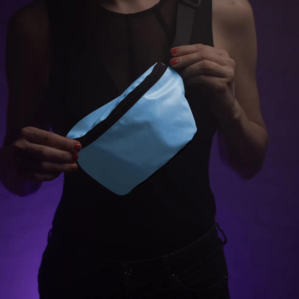 reflektierende Gürteltasche/fanny pack/Pouch in Farbvariante „Blue Curacao“ wird von einer Person am Oberkörper getragen. Sie hält den Reißverschluss der Fronttasche zwischen den Fingern. Arrangement von REFLECTIVE Berlin.