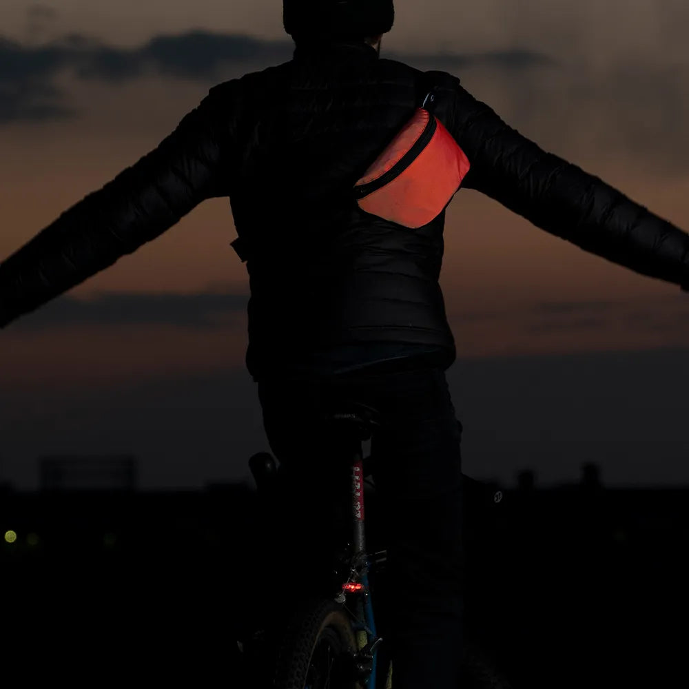 reflektierende Gürteltasche/fanny pack/Pouch in Farbvariante „Tomato red“ wird von einer Person in Rückenansicht getragen, die freihändig auf einem Fahrrad fährt. Das Foto ist bei Dämmerung mit Blitzlicht gemacht, um die rot leuchtende Reflexion hervorzuheben. Arrangement von REFLECTIVE Berlin.