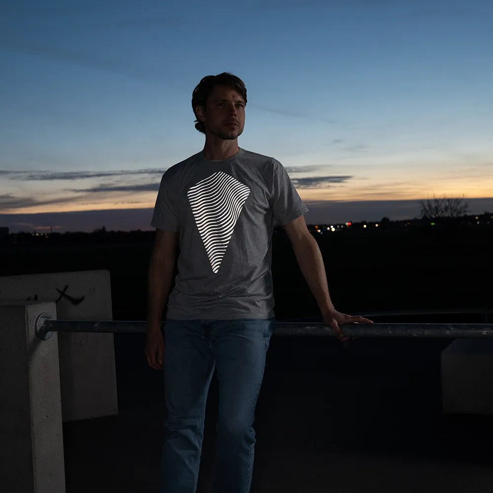 Person lehnt an einer Metallstange, trägt das reflektierende T-Shirt Dune in Grau von REFLECTIVE Berlin bei Dämmerung. Das gedruckte Design auf dem T-Shirt reflektiert weiß im Blitzlicht.