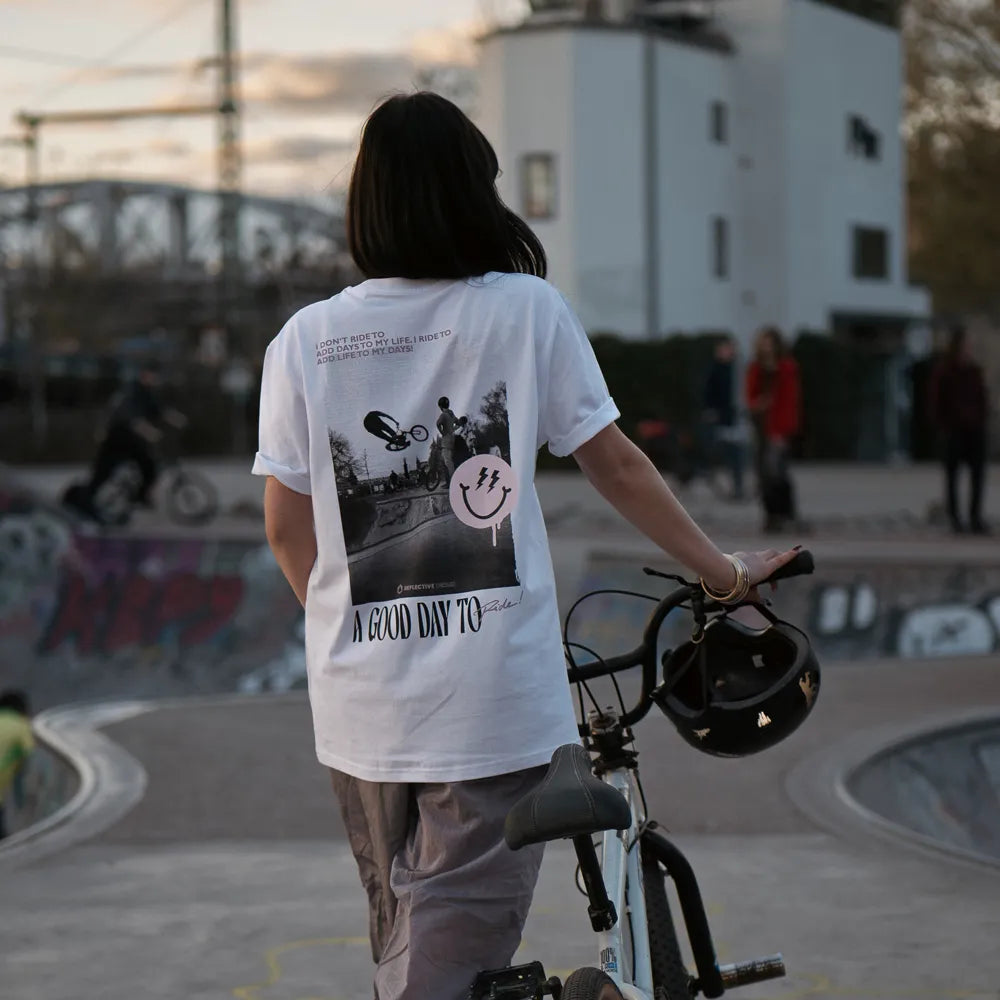 Person schiebt ein BMX Fahrrad und trägt das T-Shirt Streetlife in Weiß von REFLECTIVE Berlin mit dem Rückendruck „A Good Day to Ride“ in einem Skatepark bei Tageslicht.