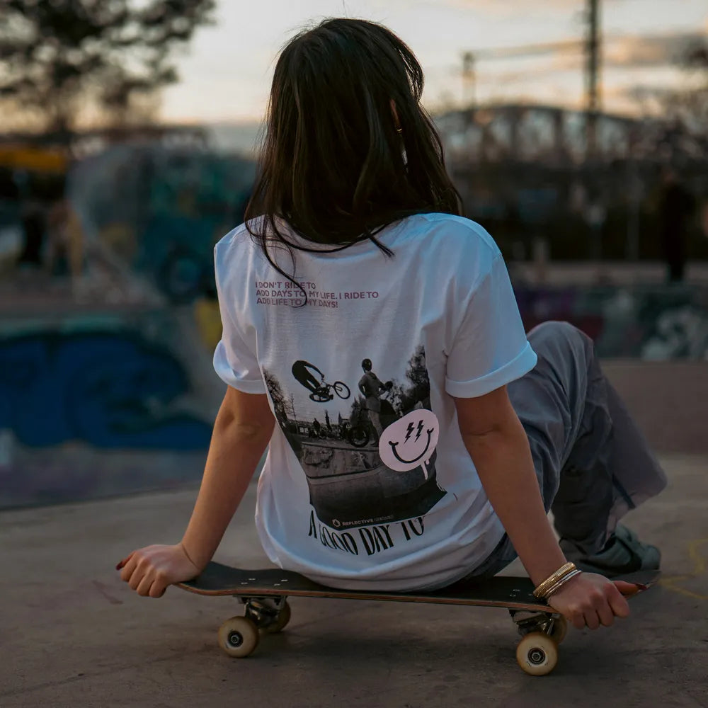 Person sitzt auf einem Skateboard und trägt das T-Shirt Streetlife in Weiß von REFLECTIVE Berlin mit dem Rückendruck „A Good Day to Ride“ in einem Skatepark bei Tageslicht.