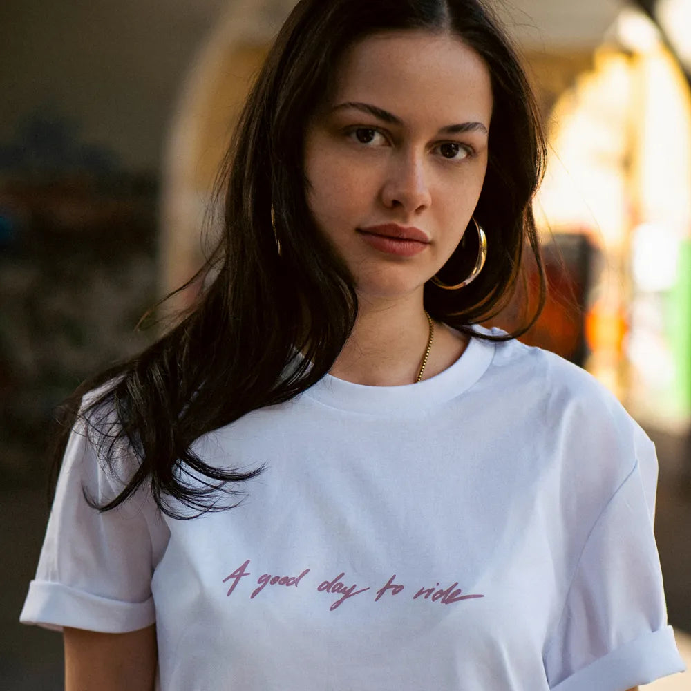 Person schaut in die Kamera und trägt das T-Shirt Streetlife in Weiß von REFLECTIVE Berlin mit dem Frontaufdruck „A Good Day to Ride“ in einem Skatepark bei Tageslicht.
