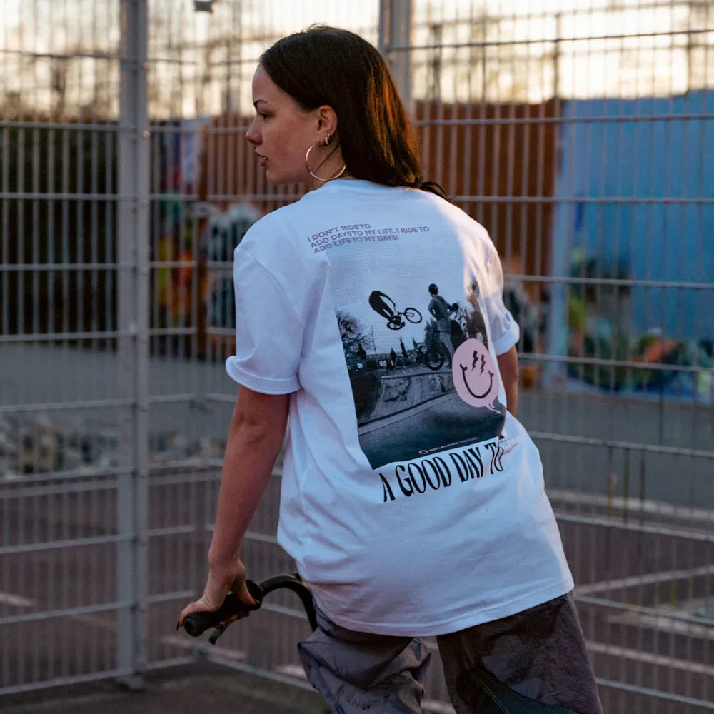 Person in Rückenansicht auf einem BMX Bike trägt das T-Shirt Streetlife in Weiß von REFLECTIVE Berlin mit dem Rückendruck „A Good Day to Ride“ in einem Skatepark bei Tageslicht.