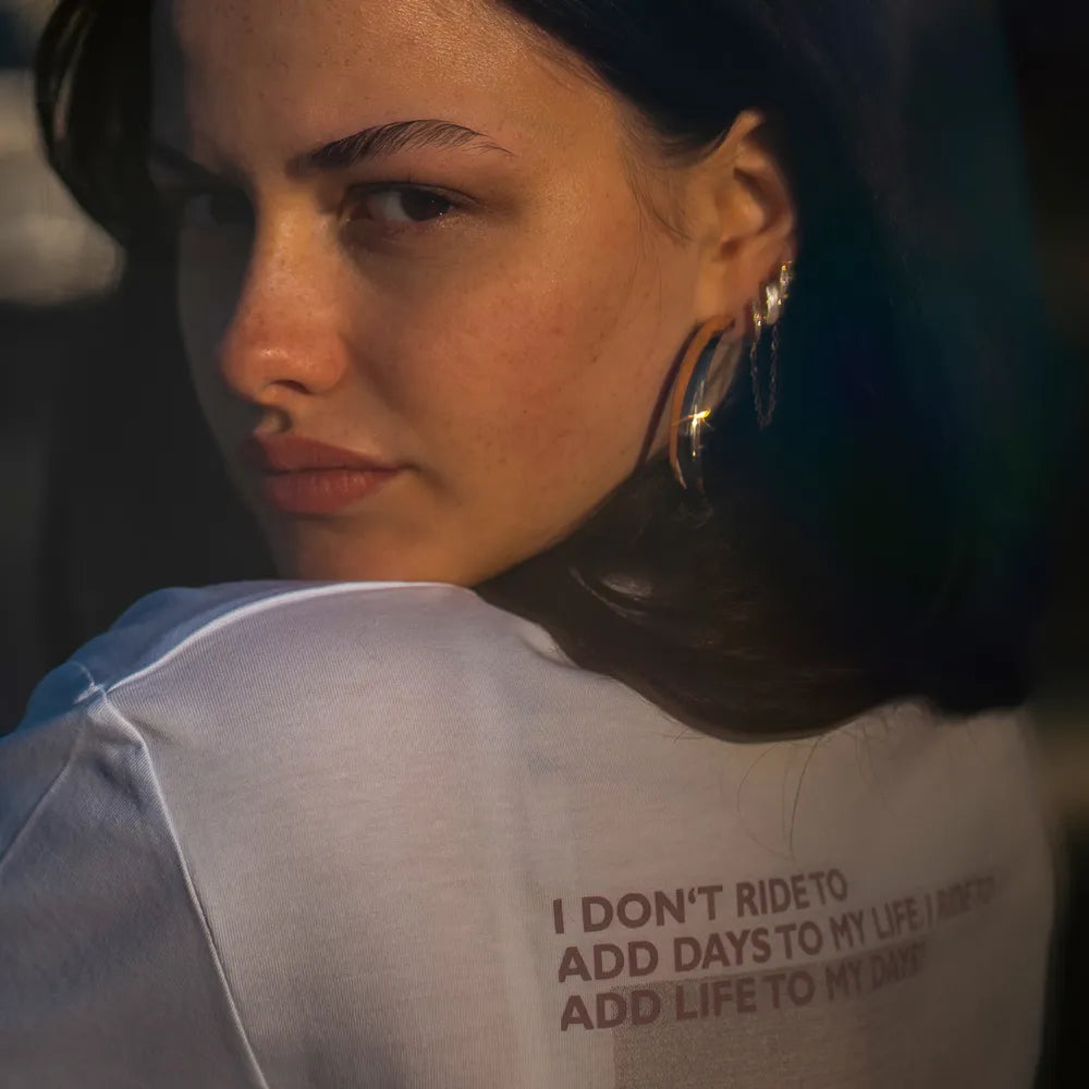 Person schaut über die Schulter nach hinten und trägt das T-Shirt Streetlife in Weiß von REFLECTIVE Berlin mit dem Rückendruck Detail „I don’t ride to add days to my life, I ride to add life to my days“