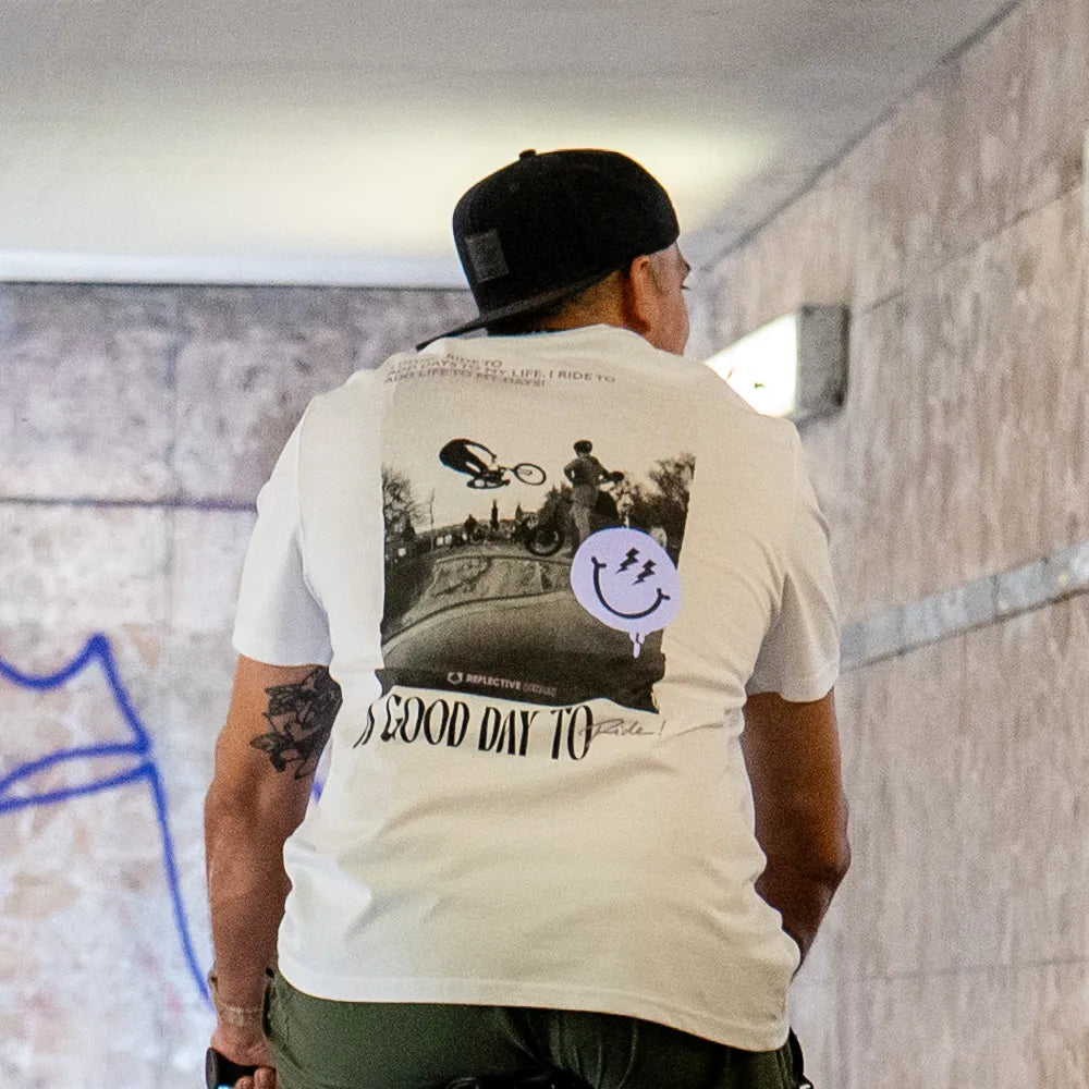 Person trägt eine umgedrehte Cap und das T-Shirt Streetlife in Weiß von REFLECTIVE Berlin mit dem Rückendruck „A Good Day to Ride“ in und sitzt auf einem Fahrrad.