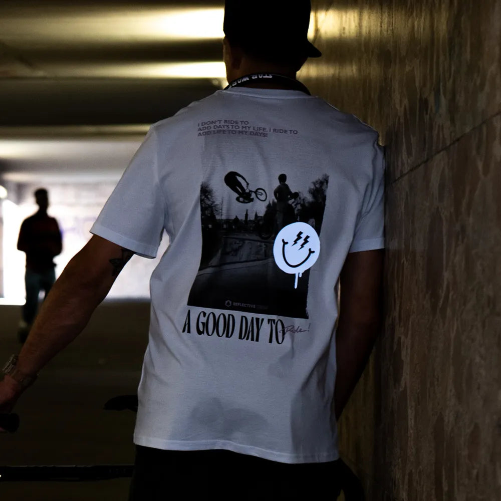 Person steht in einer Bahnhof Unterführung und rägt das T-Shirt Streetlife in Weiß von REFLECTIVE Berlin mit dem Rückendruck „A Good Day to Ride“. Die Smiley Applikation reflektiert stark.