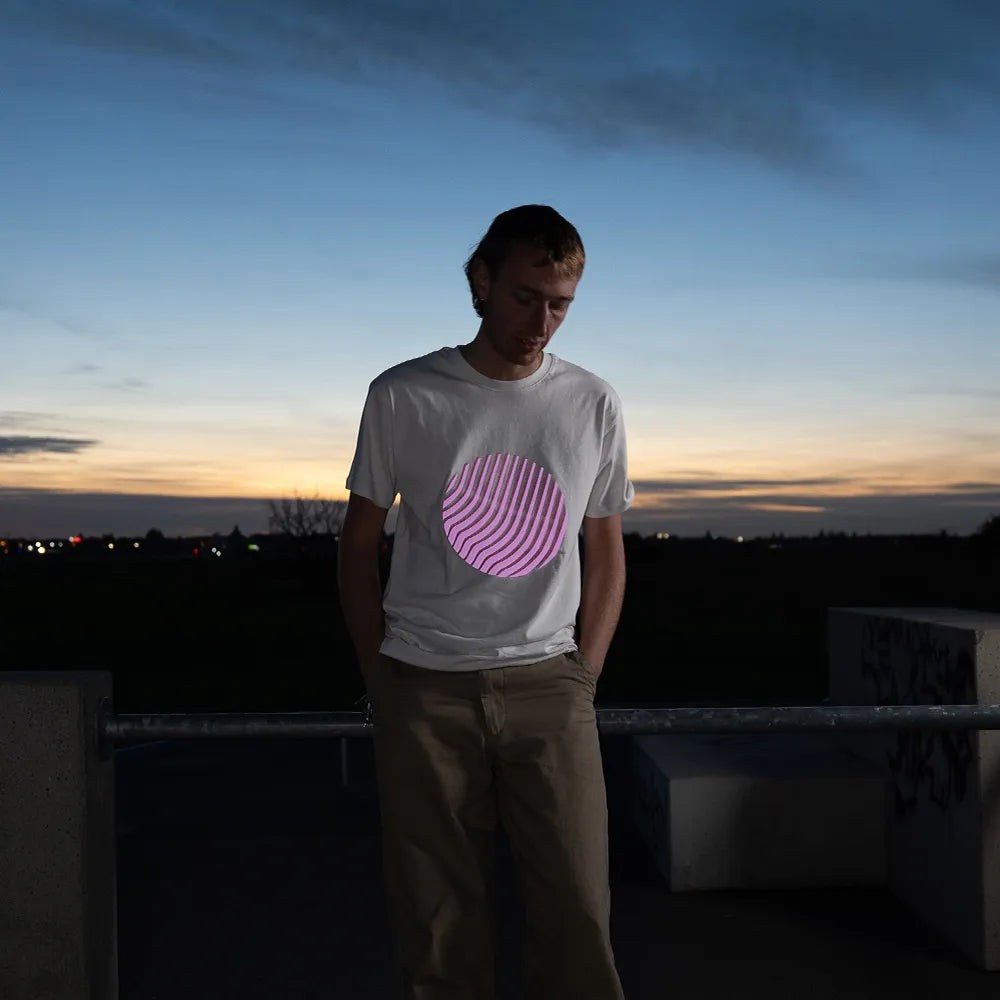 Reflektierendes T-Shirt Sunset in der Farbvariante Off-white wird von einer Person bei Dämmerung getragen. Der runde reflektierende Print auf dem T-Shirt von REFLECTIVE Berlin reflektiert in rosa.