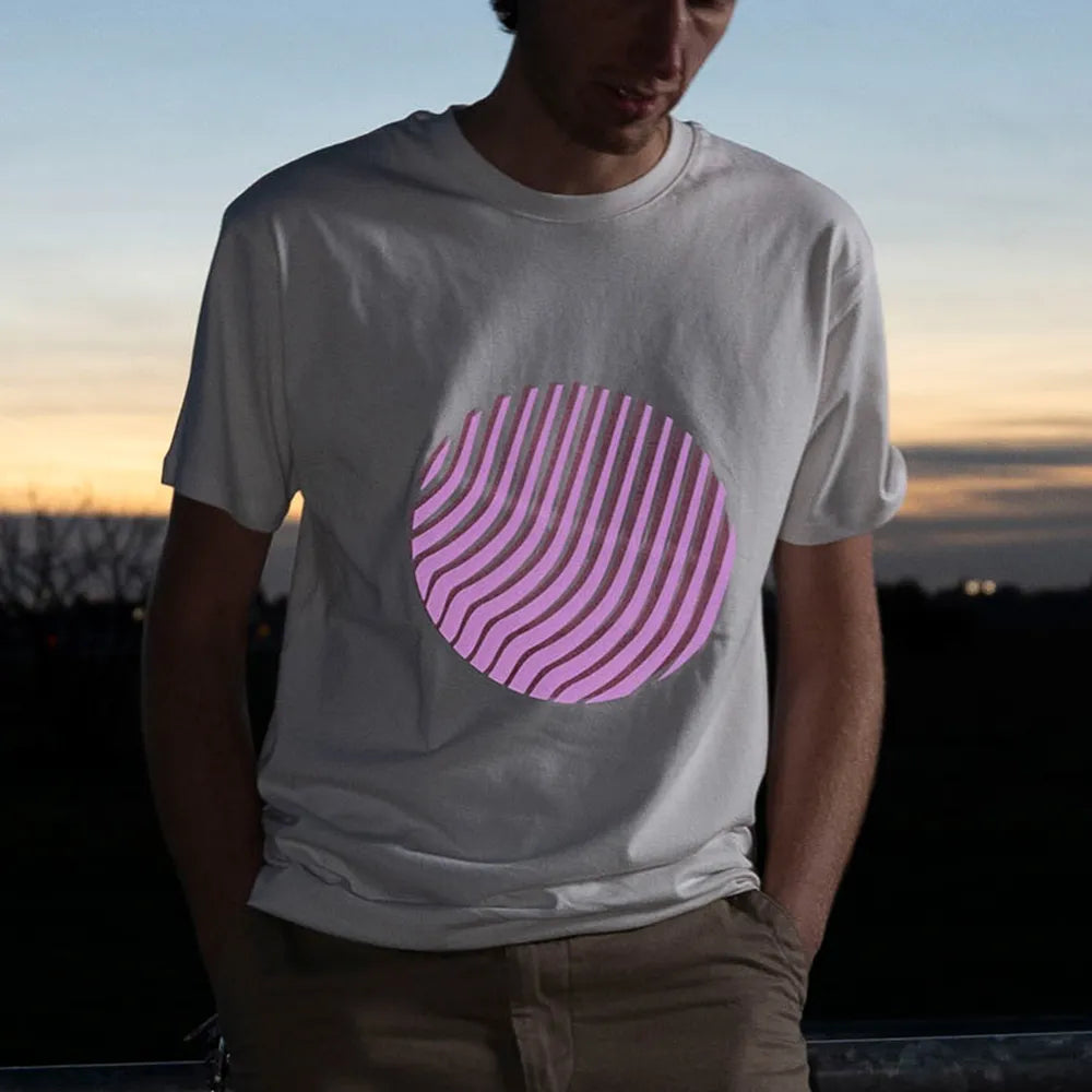 Nahaufnahme vom Reflektierenden T-Shirt Sunset in der Farbvariante Off-white. Das T-Shirt wird von einer Person bei Dämmerung getragen. Der runde reflektierende Print auf dem T-Shirt von REFLECTIVE Berlin reflektiert in rosa.