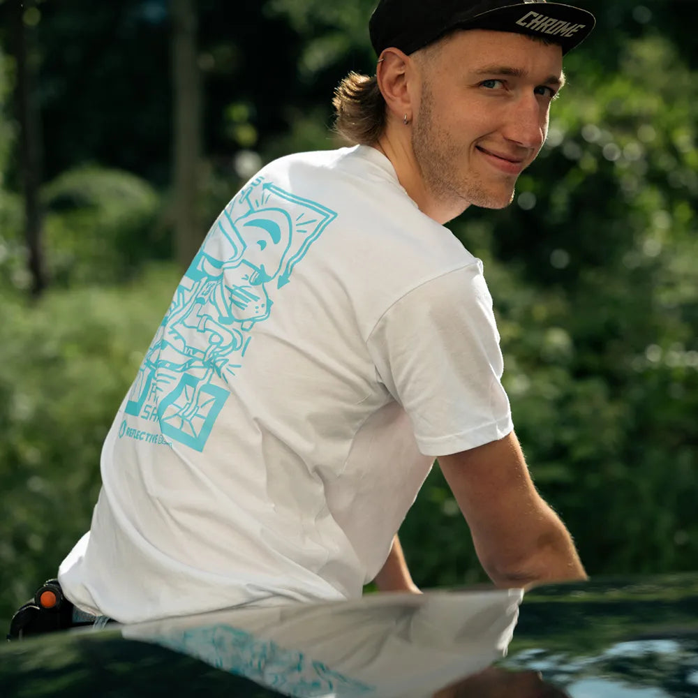 Person mit Chrome Fahrradcap in Seitenansicht Person sitzt auf einem Rennrad und trägt das Ride Reflective x VELOBerlin T-Shirt in Weiß mit blauem reflektierendem Hasen-Print von REFLECTIVE Berlin in einer Stadtszene in bei Tageslicht.