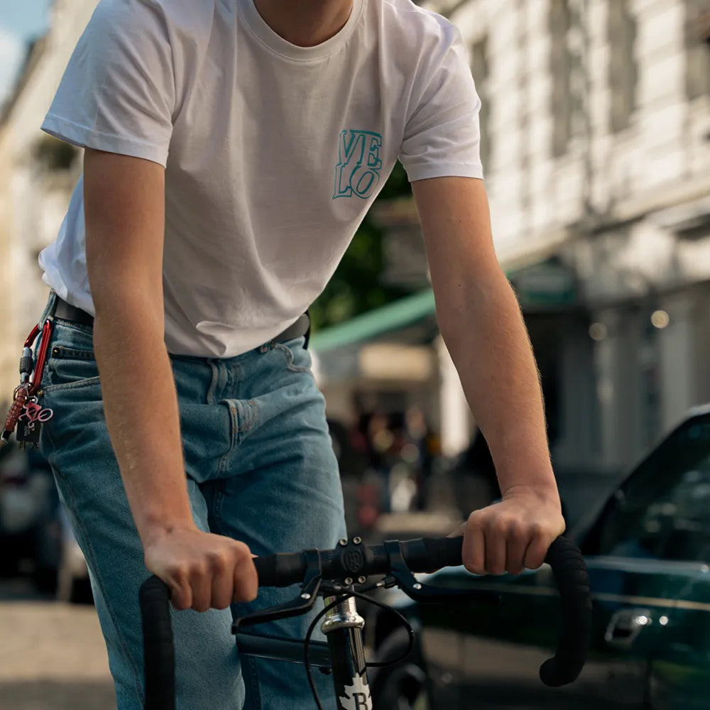 Detailausschnitt Person sitzt auf Rennrad und trägt das Ride Reflective x VELOBerlin T-Shirt von REFLECTIVE Berlin in Weiß mit blauem reflektierendem „VELO“ Logo in einer Stadtszene in bei Tageslicht.
