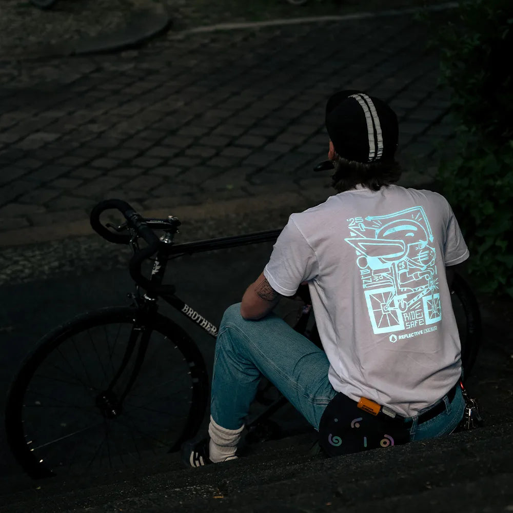 Person in Rückenansicht sitzt auf Stufen hinter einem Rennrad und trägt das Ride Reflective x VELOBerlin T-Shirt in Weiß mit blauem reflektierendem Hasen-Print von REFLECTIVE Berlin in einer Stadtszene in der Dämmerung. Der Hasen Print leuchtet und reflektiert stark.