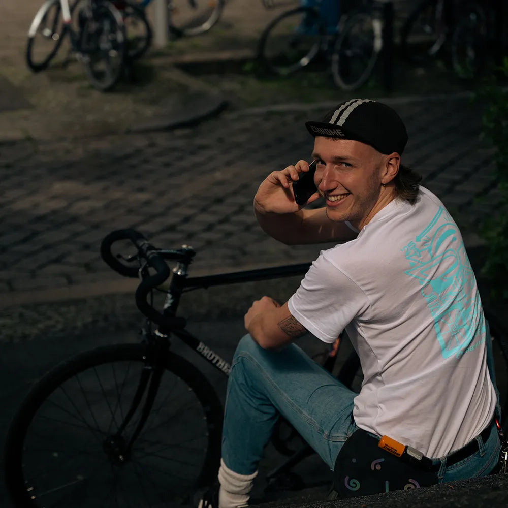 Person sitzt auf Treppe hinter einem Rennrad und schaut über die Schulter, während sie telefoniert. Person trägt das Ride Reflective x VeloBerlin T-Shirt in weiß mit blauen reflektierendem HasenPrint von REFLECTIVE Berlin in einer Stadtszene bei Tageslicht.