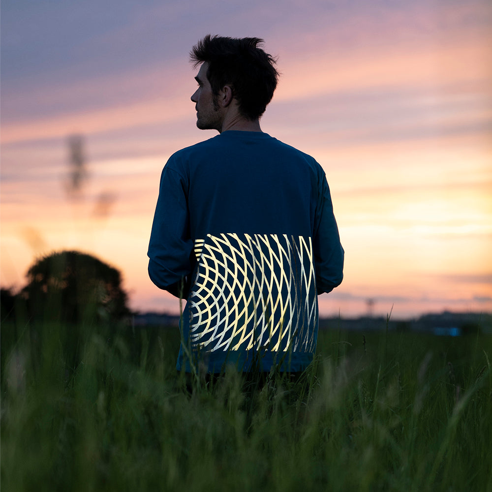 dunkelhaariger Mann in hohem Gras schaut nach links, er trägt ein Longsleeve mit reflektierendem Druck