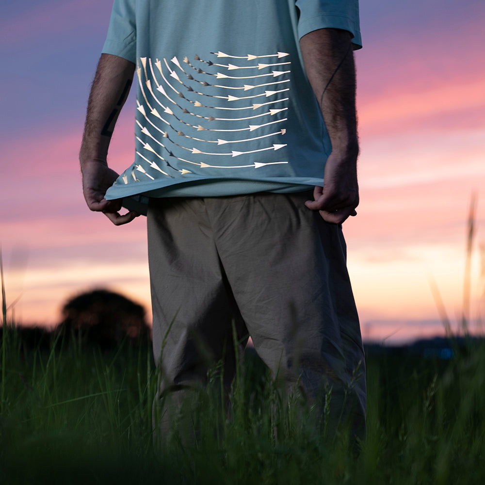 Mann auf Wiese greift mit seinen Händen an Unterkante vom T-Shirt, im Hintergrund bunter Himmel