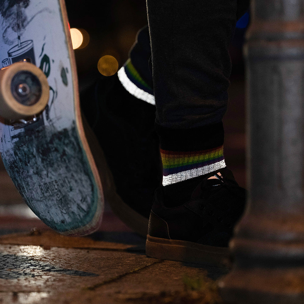 schwarze Socken mit Streifen in Regenbogen Optik und hoch reflektierendem Streifen, der von Füßen in schwarzen Turnschuhen getragen wird. die Füße stehen neben einem Skateboard. Das Arrangement von REFLECTIVE Berlin ist bei Dämmerung mit Stadtlichtern im Hintergrund aufgenommen, um die Reflexion des Streifens hervorzuheben.