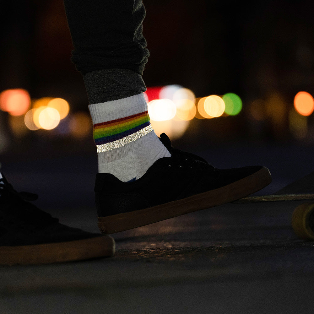 weißer Socken mit Streifen in Regenbogen Optik und hoch reflektierendem Streifen, der von einem Fuß in schwarzen Turnschuh getragen wird. Das Arrangement von REFLECTIVE Berlin ist bei Dämmerung mit Stadtlichtern im Hintergrund aufgenommen, um die Reflexion des Streifens hervorzuheben.