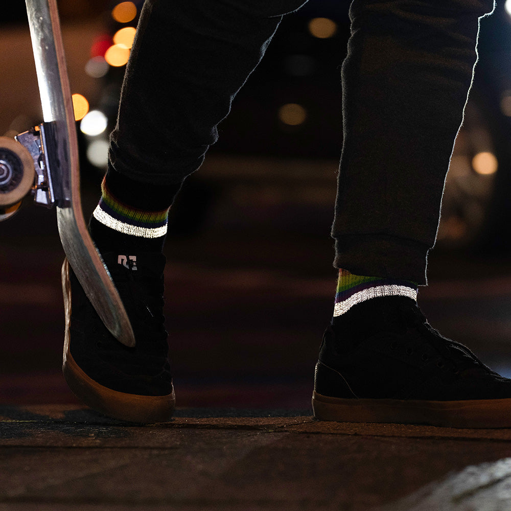 schwarze Socken mit Streifen in Regenbogen Optik und hoch reflektierendem Streifen, die von Füßen in schwarzen Turnschuhen getragen werden. Die Füße stehen neben einem aufgerichteten Skateboard. Das Arrangement von REFLECTIVE Berlin ist bei Dämmerung mit Stadtlichtern im Hintergrund aufgenommen, um die Reflexion des Streifens hervorzuheben.