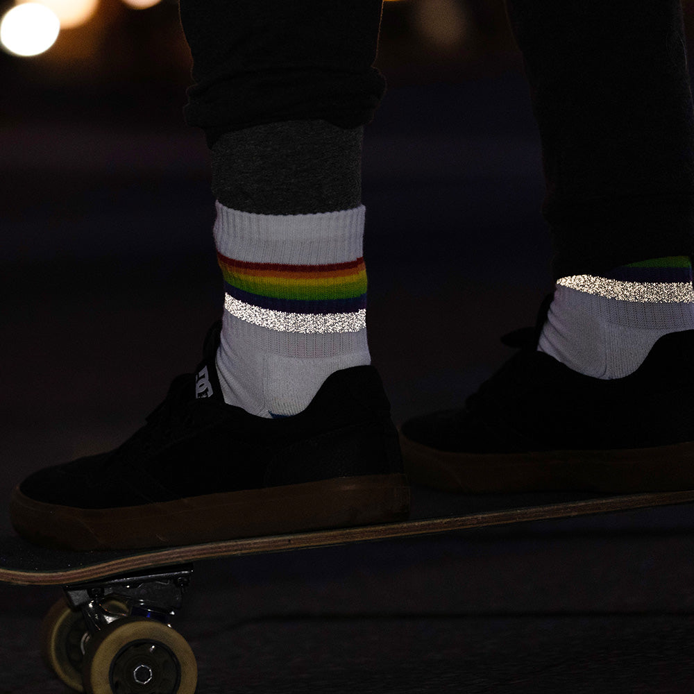 weiße Socken mit Streifen in Regenbogen Optik und hoch reflektierendem Streifen, die von zwei Füßen in schwarzen Schuhen getragen werden. Die Füße stehen auf einem Skateboard. Das Arrangement von REFLECTIVE Berlin ist bei Dämmerung aufgenommen, um die Reflexion hervorzuheben.