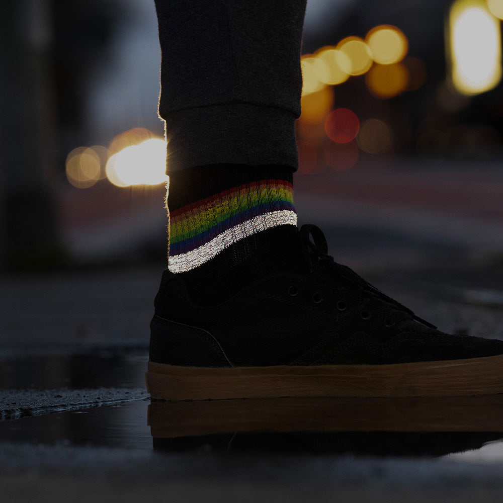 schwarzer Socken mit Streifen in Regenbogen Optik und hoch reflektierendem Streifen, der von einem Fuß in schwarzen Turnschuh getragen wird. Das Arrangement von REFLECTIVE Berlin ist bei Dämmerung mit Stadtlichtern im Hintergrund aufgenommen, um die Reflexion des Streifens hervorzuheben.