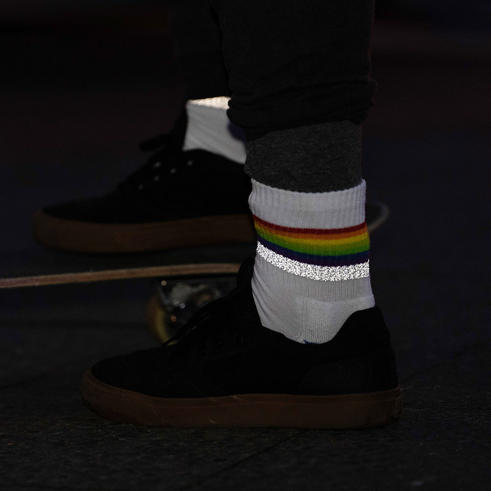 reflektierende weiße Socken in Farbvariante „weiß Regenbogen“ mit Streifen in Regenbogen Optik und hoch reflektierendem Streifen an einem Fuß der vor einem Skateboard steht. Die Socken von REFLECTIVE Berlin reflektieren stark in der Dunkelheit.