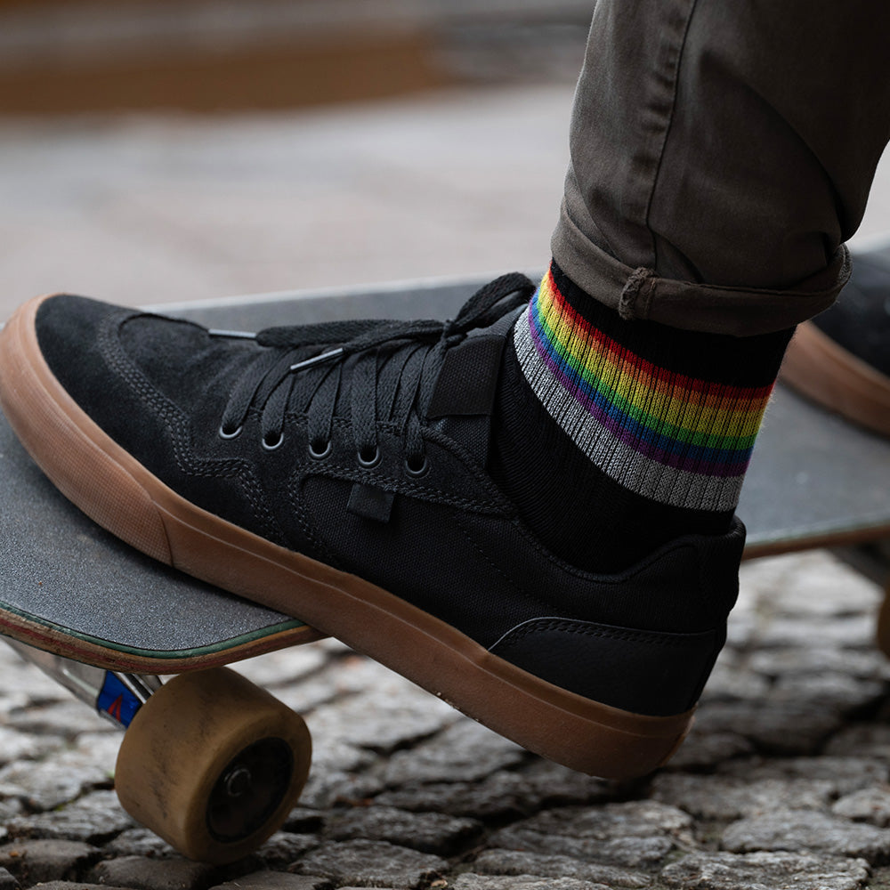 schwarze Socken mit Streifen in Regenbogen Optik und hoch reflektierendem Streifen, der von Füßen in schwarzen Turnschuhen getragen wird. Die Füße stehen auf einem Skateboard. Das Arrangement von REFLECTIVE Berlin ist bei Tageslicht aufgenommen.