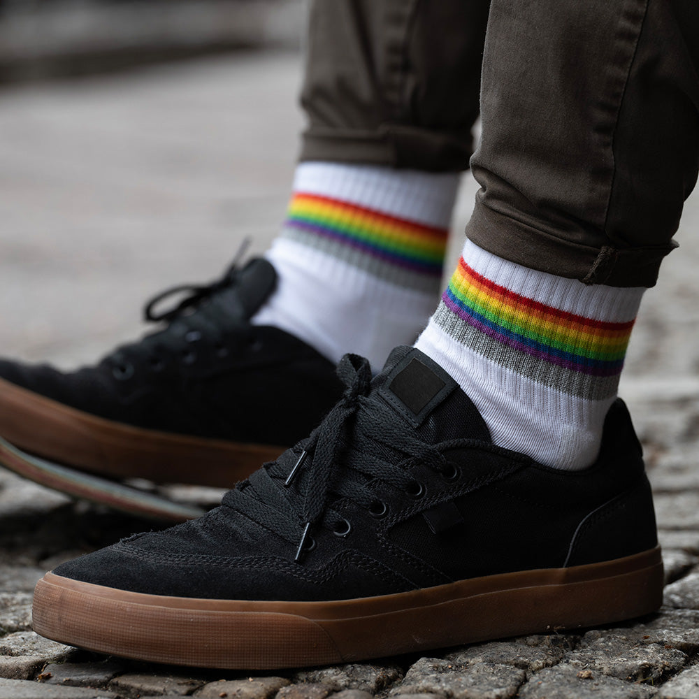 weiße Socken mit Streifen in Regenbogen Optik und hoch reflektierendem Streifen, die von zwei Füßen in schwarzen Schuhen getragen werden. Das Arrangement von REFLECTIVE Berlin ist bei Tageslicht mit einem unscharfen Hintergrund aufgenommen.