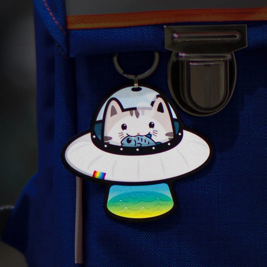 Reflektierender Anhänger ‚Reflective Hangers‘  in der Variante ‚Space Cat’. Ein Reflektor Anhänger der eine Katze in einem Ufo zeigt. Die Katze hat einen Fisch im Mund. Der stark reflektierende Anhänger hängt an einem blauen Schulrucksack von REFLECTIVE Berlin.