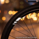 Reflektierender Aufkleber „Racer“ in Farbvariante „schwarz“ in Größe 20mm auf schwarzer Fahrrad Felge im Dunkeln mit verschwommenen Lichtern im Hintergrund. von REFLECTIVE Berlin.