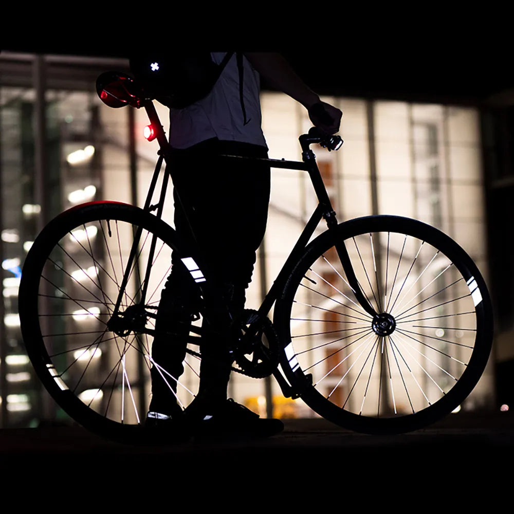 Reflektierender Felgen Aufkleber „Racer“ in Farbvariante „weiß“ in Größe 40mm auf schwarzem Fahrrad Felgen im Dunkeln mit verschwommenen Lichtern im Hintergrund. von REFLECTIVE Berlin. 