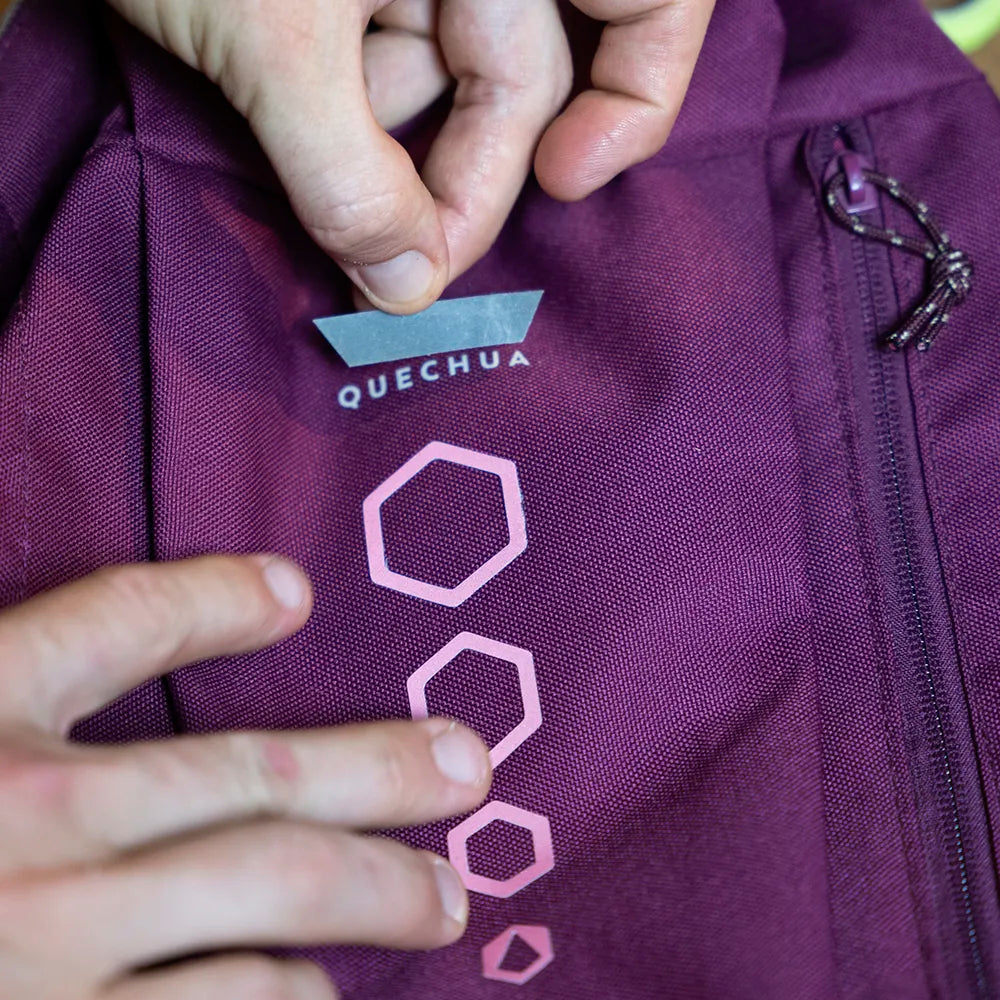 Reflektierende Aufkleber Textil Stoff „DIY“ in Farbvariante „weiß“, zugeschnitten in einen Streifen um ein QUECHUA Logo auf einem lila Rucksack abzudecken. Sticker von REFLECTIVE Berlin.
